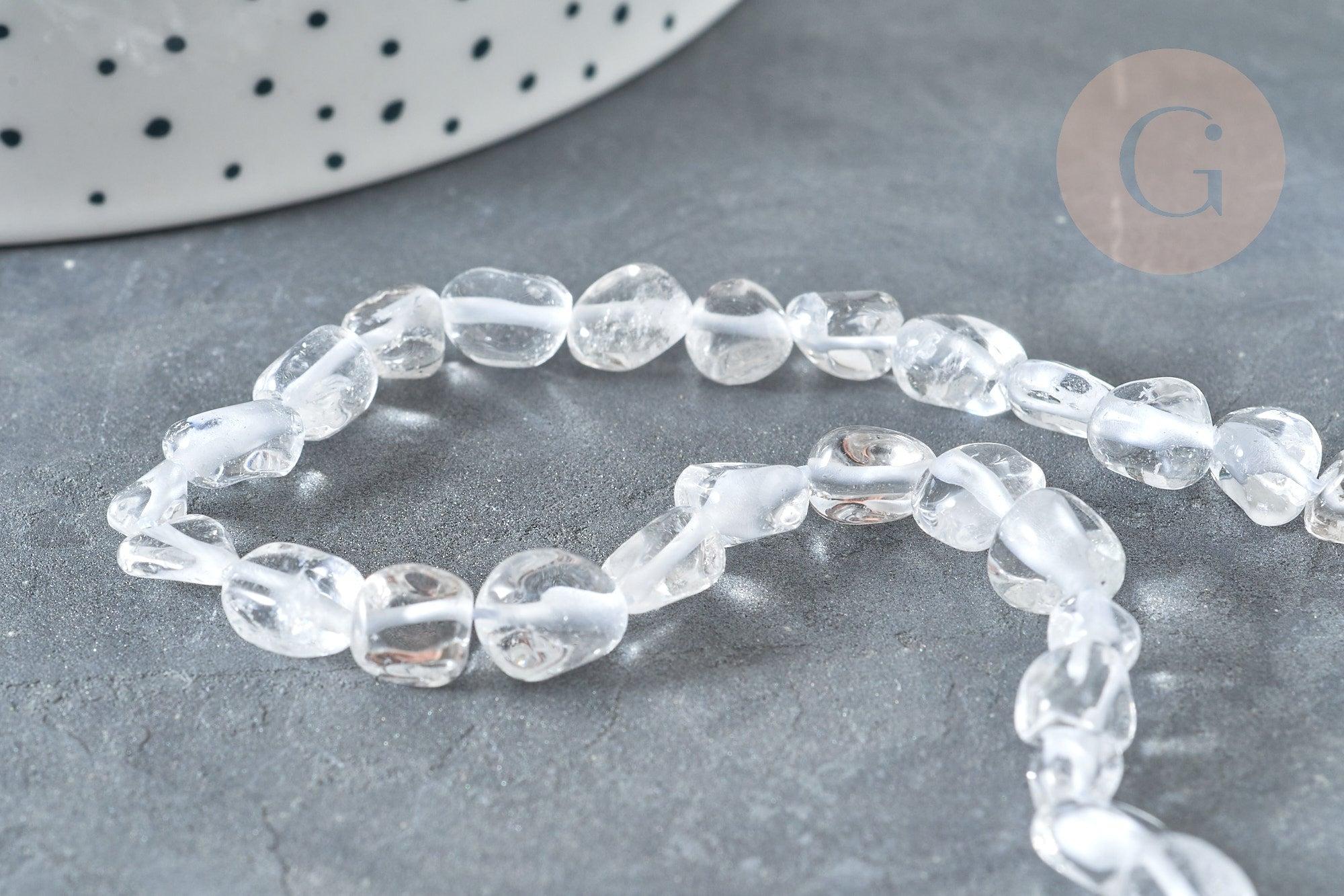 Close-up of Perles cristal de roche roulée, featuring clear, irregular-shaped quartz beads, laid on a gray surface with a dotted white container partially visible.