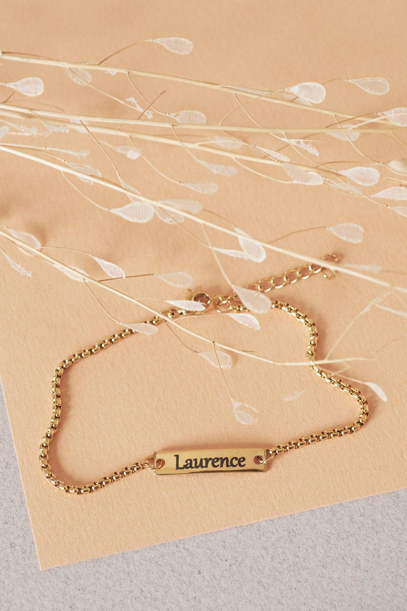Bracelet chaîne doré avec plaque gravée "Laurence", sur fond beige clair avec herbes séchées.
